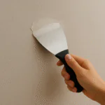 Mão aplicando massa corrida com espátula em parede, mostrando como nivelar e preparar a superfície para pintura